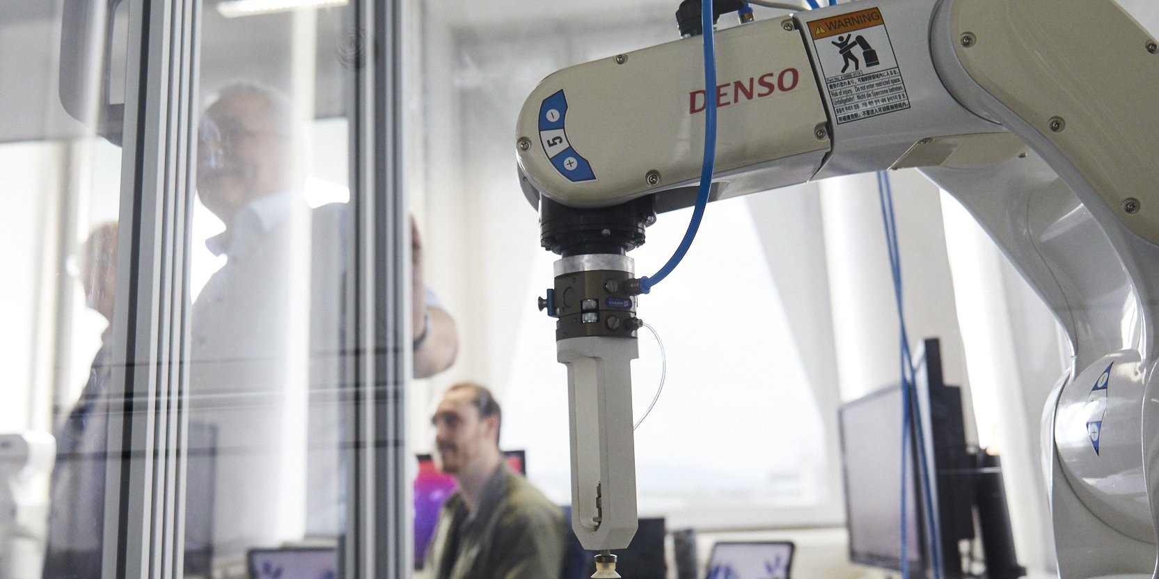 Robo-Greifarm in einem Glasgehäuser, dahinter verschwommen Menschen im Labor