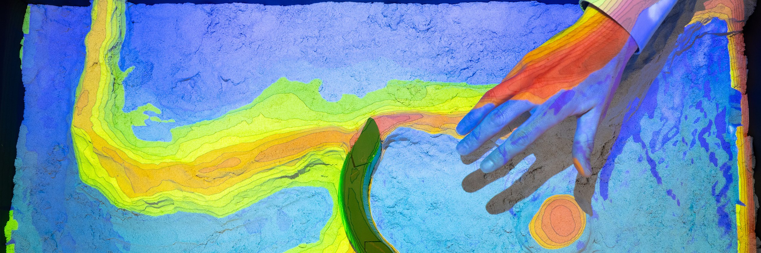 Eine Hand zeigt in die Sandbox, die in Grün-, Blau- und Brauntönen eine Landschaft abbildet. 