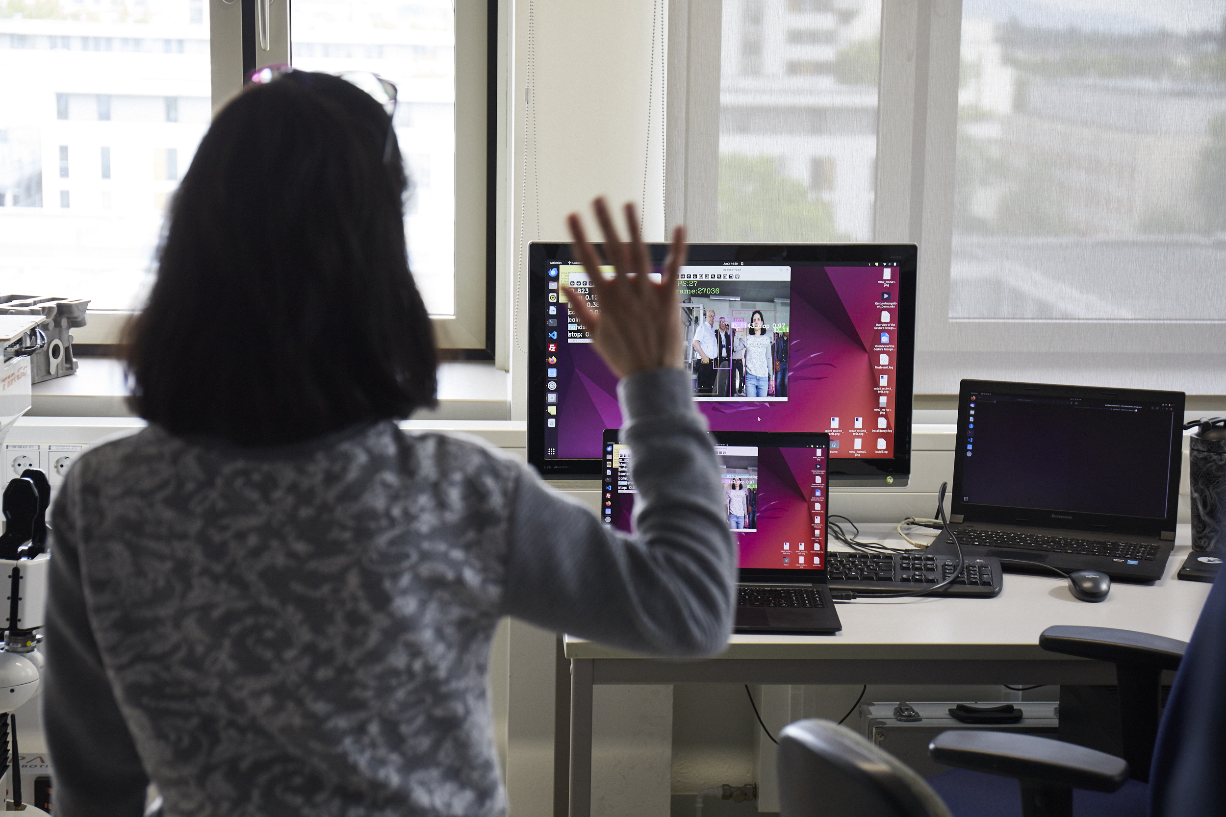 Wissenschaftlerin mit dem Rücken zur Kamera winkt, ihr Bild wird vorne auf einen Laptop projiziert